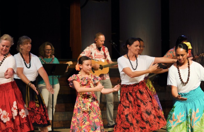Several women and a little girl in colorful Hawaiian garb hula dance on a stage in a church.