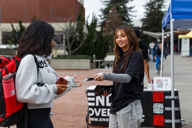 A young person smiles as they hand something to another young person in front of a table with a sign that reads "end overdose"