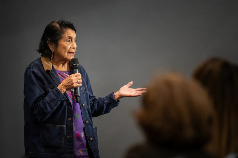 Dolores Huerta speaks into a handheld microphone, gesturing with one hand toward an audience while standing against a plain indoor backdrop.