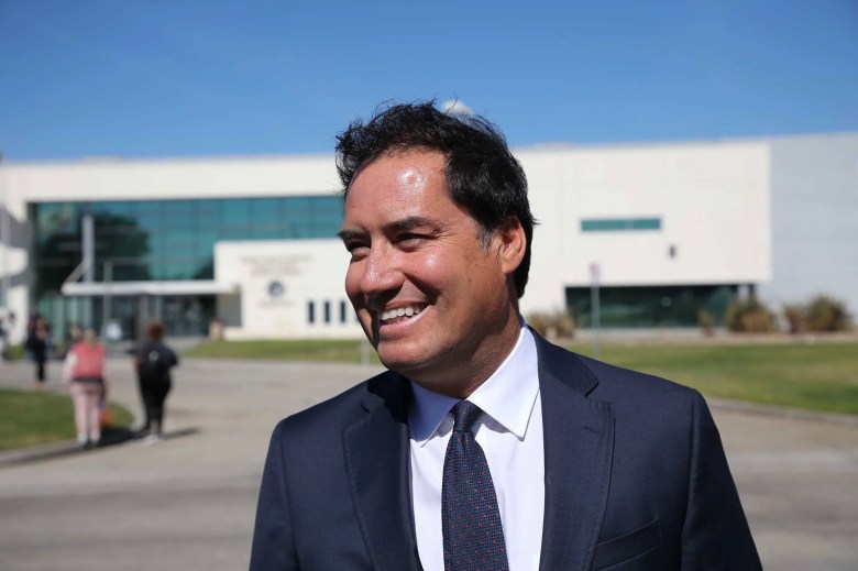 A person in a suit and tie smiles while standing outdoors in front of a modern building, with a few people walking in the background under a clear blue sky.