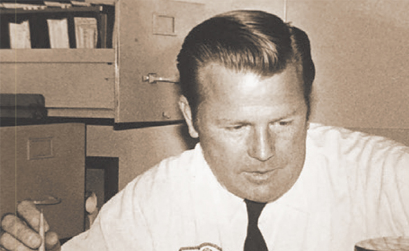 A person with neatly combed hair and a tie looks downward while seated at a desk, with filing cabinets and shelves visible behind them.