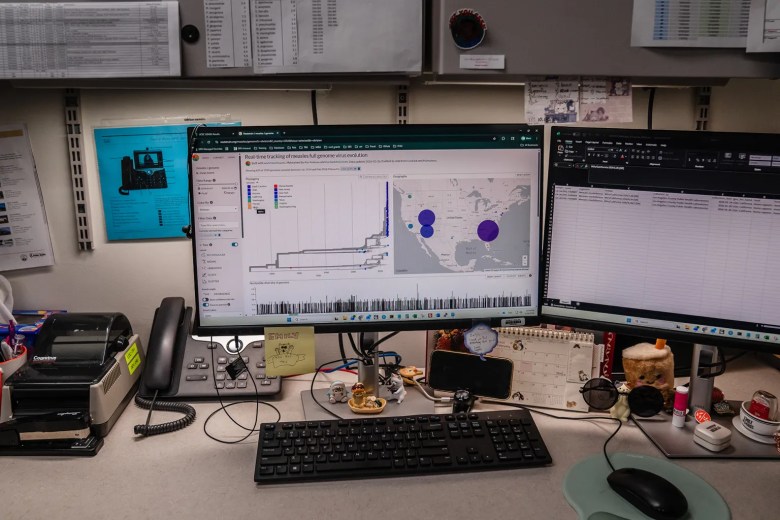 Cluttered office desk with dual monitors displaying data charts and spreadsheets.