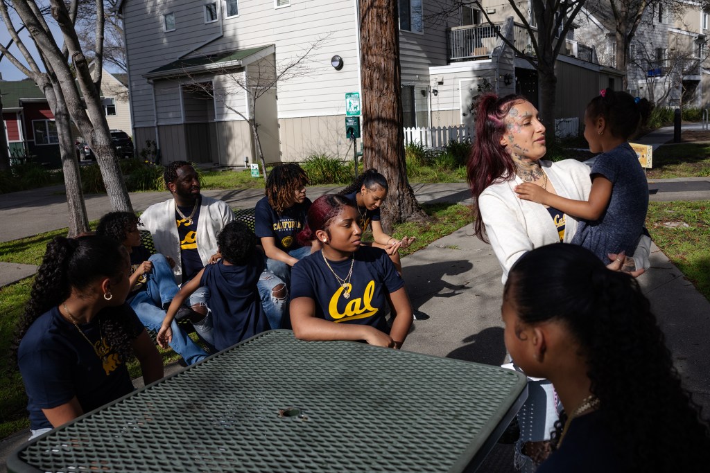 A family of eight children sits on park benches in a small park near apartment buildings, wearing matching blue shirts with the UCLA logo.