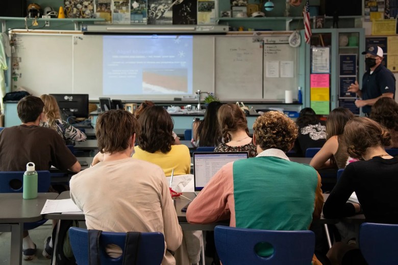 Students in a classroom facing a teacher and a projector screen.