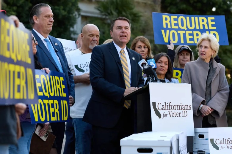 A lawmaker, wearing a black suit with a striped yellow tie, stands behind a lectern while surrounded by people holding blue and yellow signs that say "REQUIRE VOTER ID."