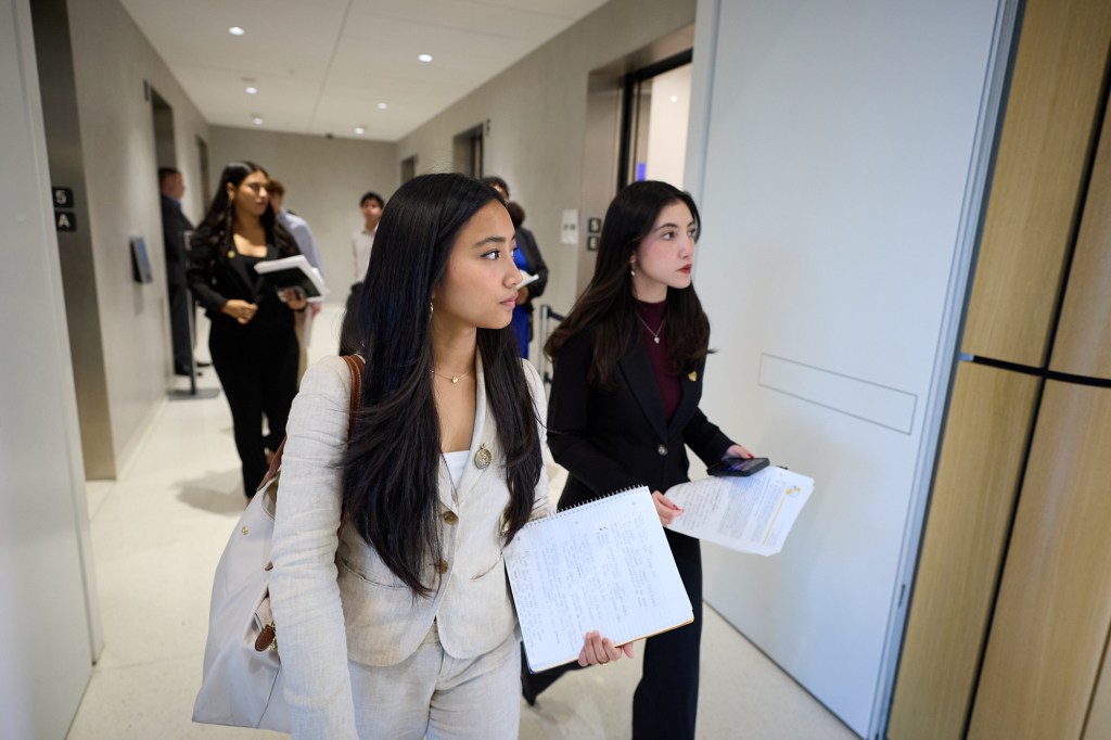 Two young adults dressed in business clothing walk down a hallway while holding notebooks.