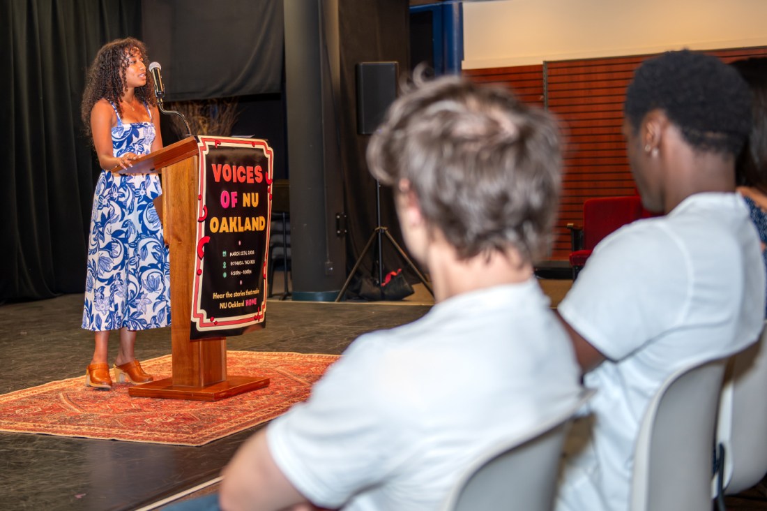 A student wearing a blue and white dress stands at a podium speaking on a staged platform doing the Voices of NU event, viewed from behind several seated audience members. 