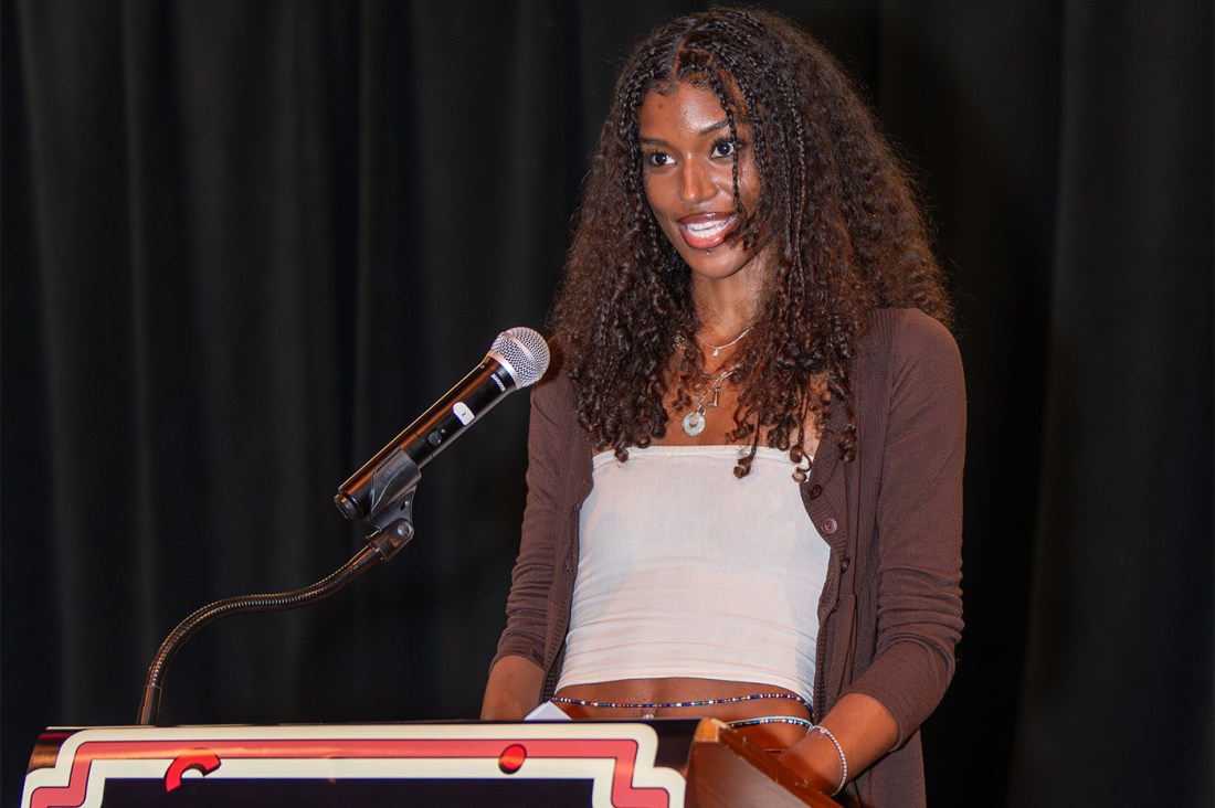 A student with dark curly hair smiles while standing at a podium with a microphone at the voices of NU event. She is wearing a white top and brown cardigan. 