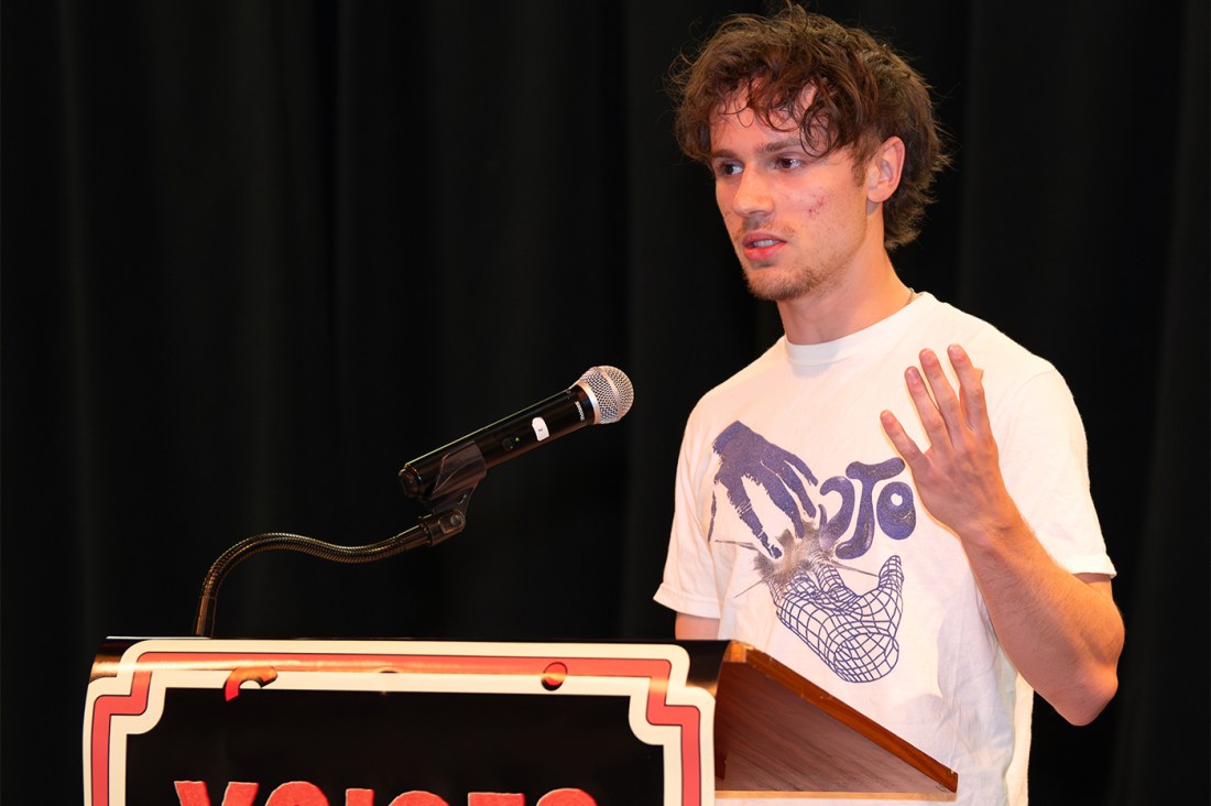 A student with curly hair wearing a white t-shirt speaks at a podium, gesturing with one hand while addressing the audience. 