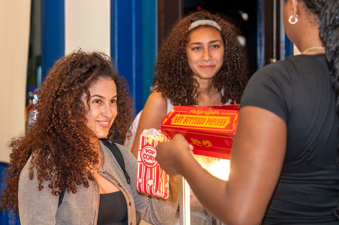 Three people in conversation with each other, one with their back to the camera. One facing the camera holds a red popcorn bucket. 