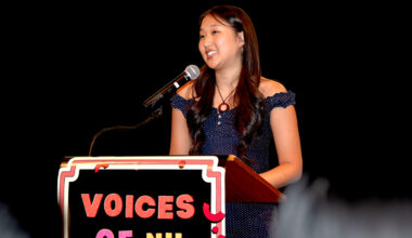 A young woman with long dark hair smiles while speaking at a wooden podium on a darkened stage at the Voices of NU event. The podium has a sign that says 'Voices of NU' on it in red and pink lettering on a black background.