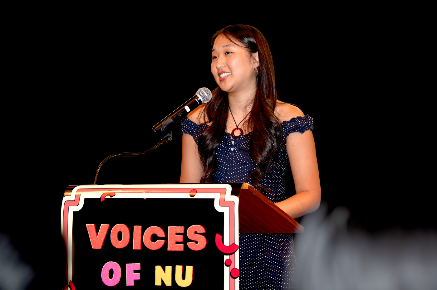 A young woman with long dark hair smiles while speaking at a wooden podium on a darkened stage at the Voices of NU event. The podium has a sign that says 'Voices of NU' on it in red and pink lettering on a black background.