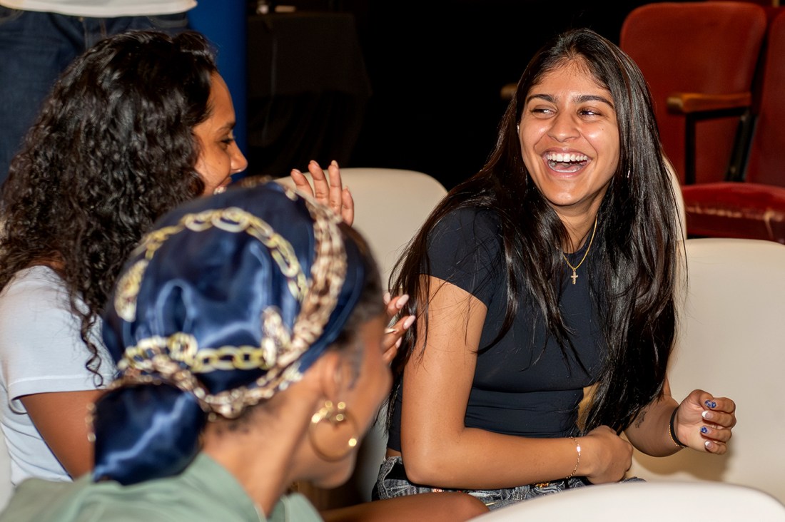 Three students laugh and chat together while seated at the Voices of NU event. The student in the foreground faces away from the camera, wearing a decorative blue and gold scarf on their head. The students next to and across from her laugh openly. 