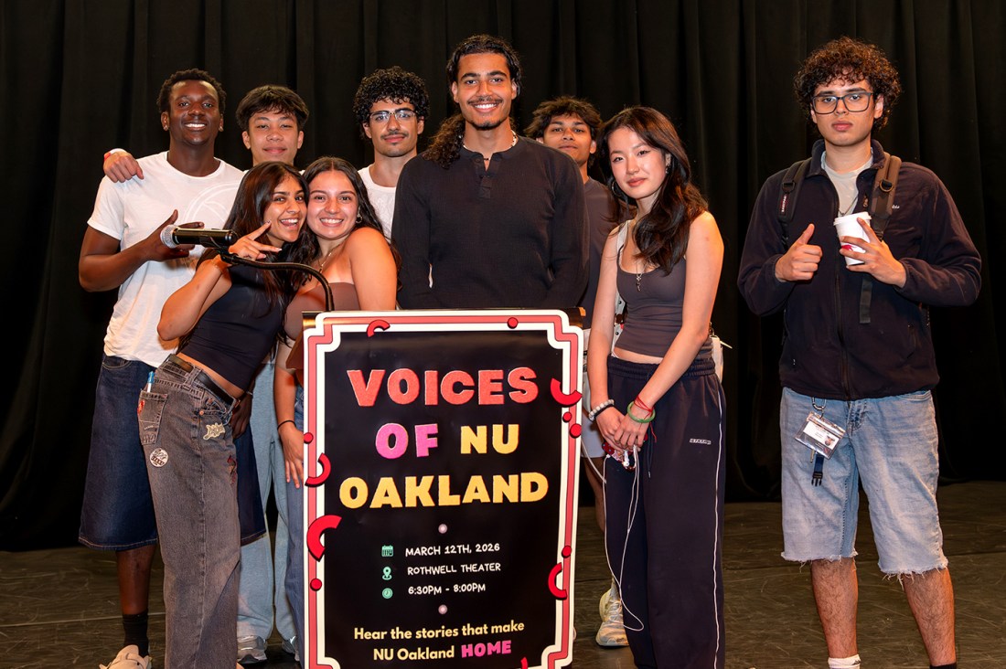 A group of nine students pose together smiling and making hand gestured in front of a podium that has a Voices of NU Oakland sign on it. 
