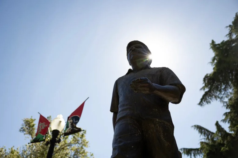 A low-angle view of a bronze statue of a person standing with one hand slightly extended, silhouetted against a bright sky. Sunlight flares from behind the figure’s head, while a streetlamp with red and green banners and leafy trees frame the scene below.