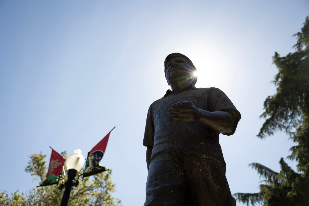 A low-angle view of a bronze statue of a person standing with one hand slightly extended, silhouetted against a bright sky. Sunlight flares from behind the figure’s head, while a streetlamp with red and green banners and leafy trees frame the scene below.