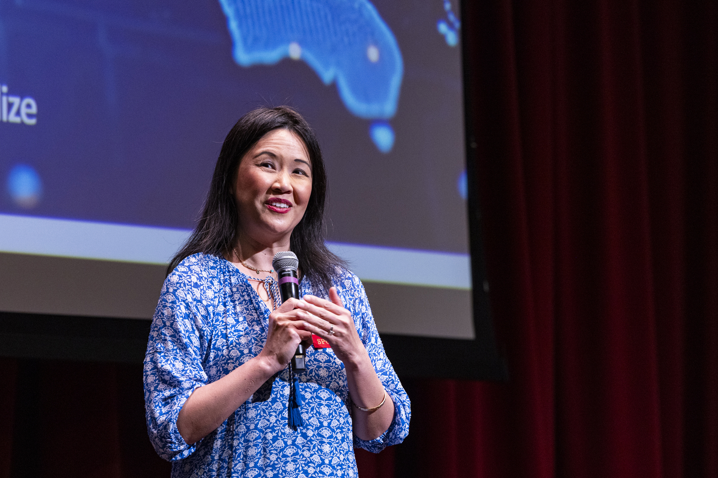 A speaker gestures while holding a microphone and speaking in front of a projected screen during the Global Health Consortium at Northeastern Oakland.