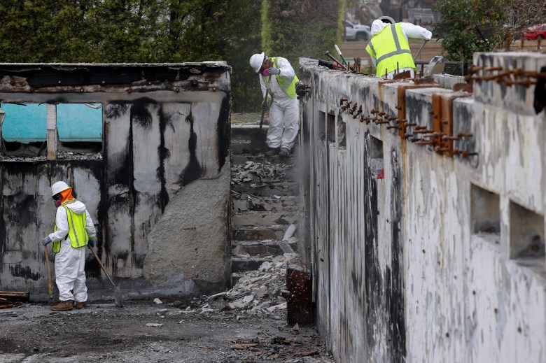 People wearing white jumpsuits and yellow safety vests gather up debris from a home destroyed during a wildfire.