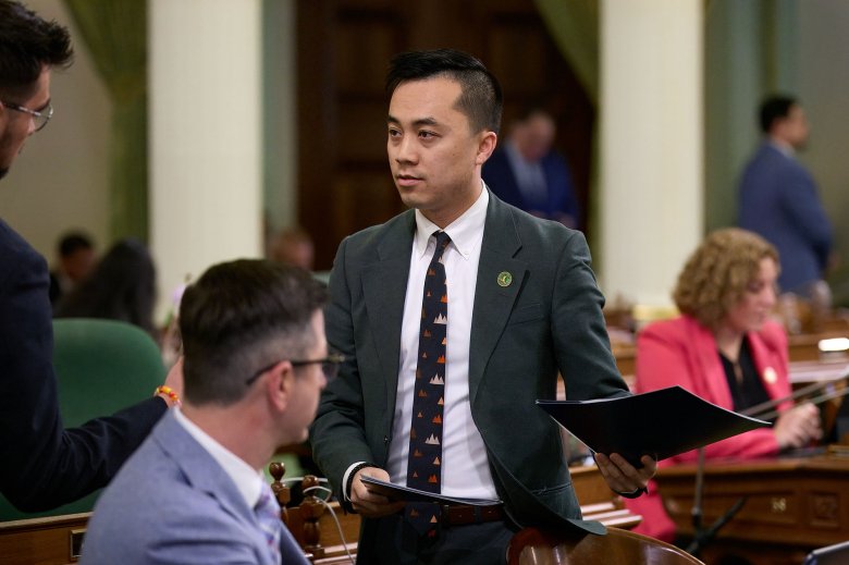 A person wearing a green suit and a patterned black tie holds folders in both hans as he talks with other lawmakers on a legislative floor room.