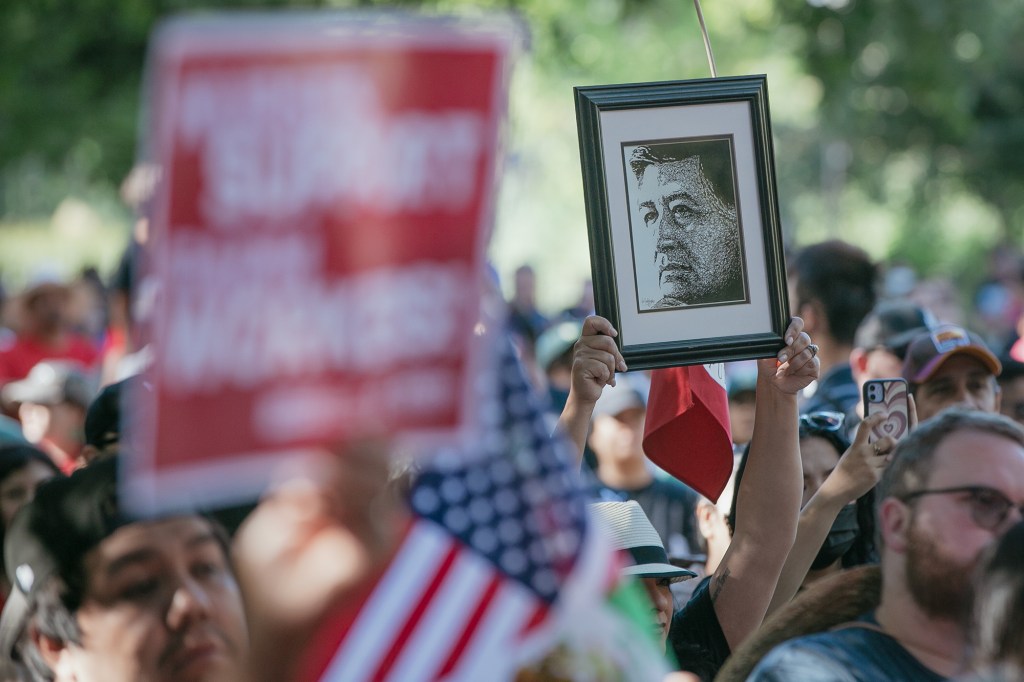 A person in a crowded outdoor gathering holds up a framed black-and-white portrait above their head. Around them, a dense group of people stands close together, some holding signs and small American flags, while others record on their phones. The background is softly blurred with trees and daylight, emphasizing the raised portrait as a focal point amid the crowd.