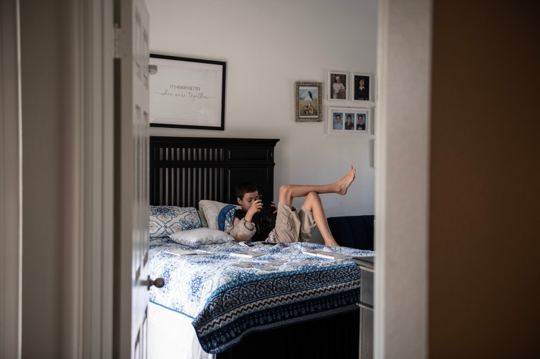 A young boy lounges on a bed with a blue and white patterned blanket, holding a camera, in a softly lit bedroom decorated with family photos and a framed sign that reads "It's always better when we're together."