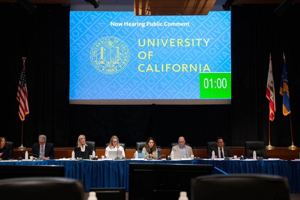 A wide view of a people sitting in front of a blue table as a large projector displays a digital blue graphic that says "University of California" with a one minute timer on it.