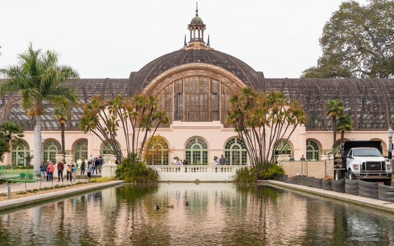 Visitors and construction works mill around the Botanical Building in Balboa Park. The color of green palms and the building's tan facade refracts off the reflecting pool, home to three ducks.