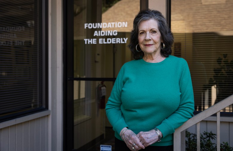 An older adult wearing a green sweater stands outside a glass door with the words “Foundation Aiding the Elderly” printed on it, hands clasped and looking toward the camera.