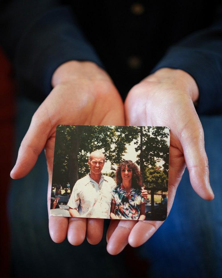 A pair of open hands holds a small printed photograph showing two people standing together outdoors beneath trees.