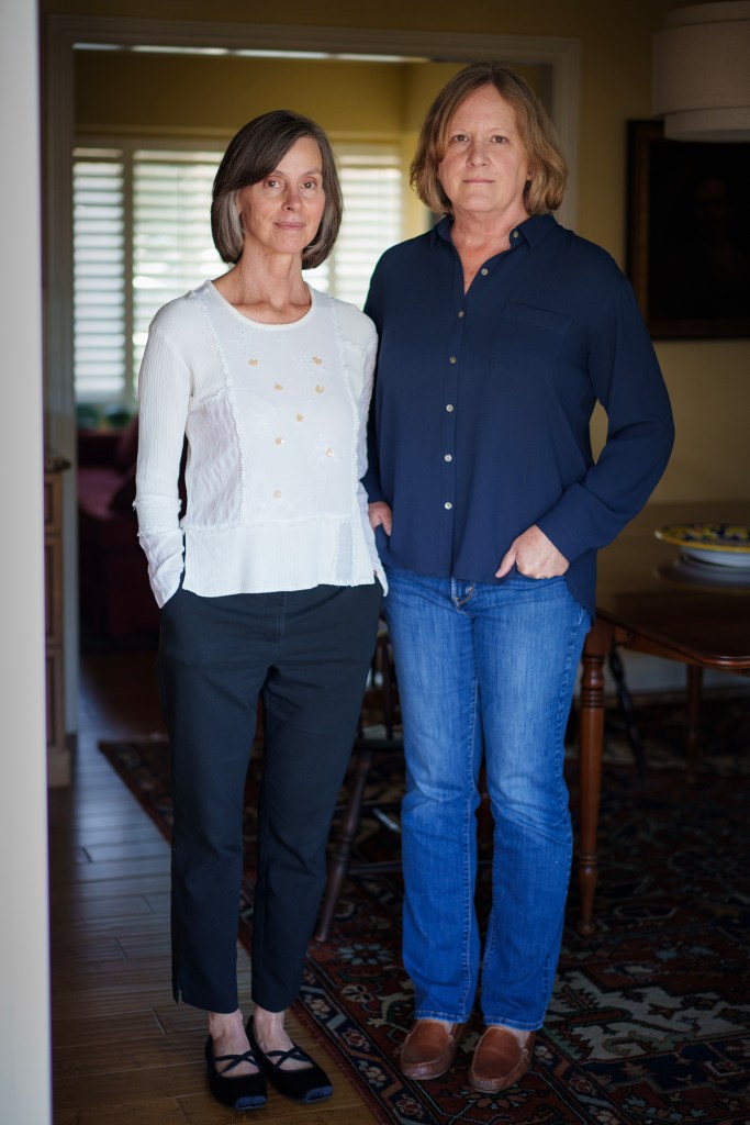Two people stand side by side indoors, facing the camera. One wears a light long-sleeve top and dark pants, and the other wears a dark button-up shirt and jeans, with hands in their pockets. A doorway with window blinds and a softly lit room appears behind them.