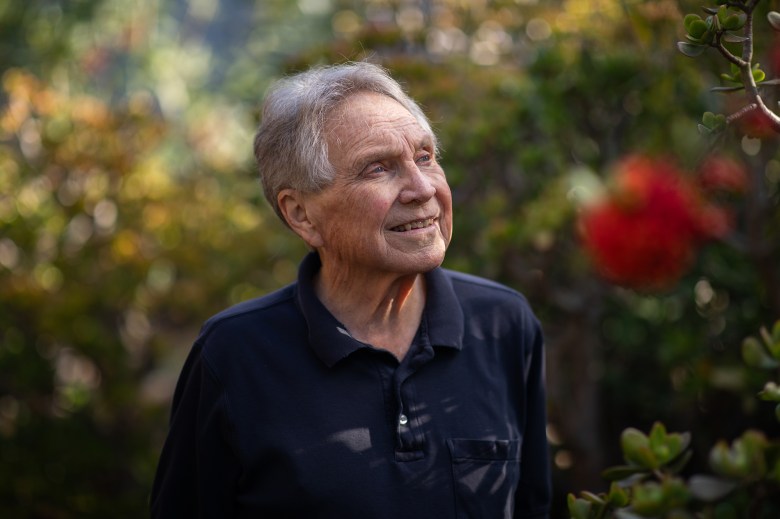An older adult with short gray hair stands outdoors in a garden, smiling slightly while looking off to the side, with soft greenery and a blurred red flower in the foreground.