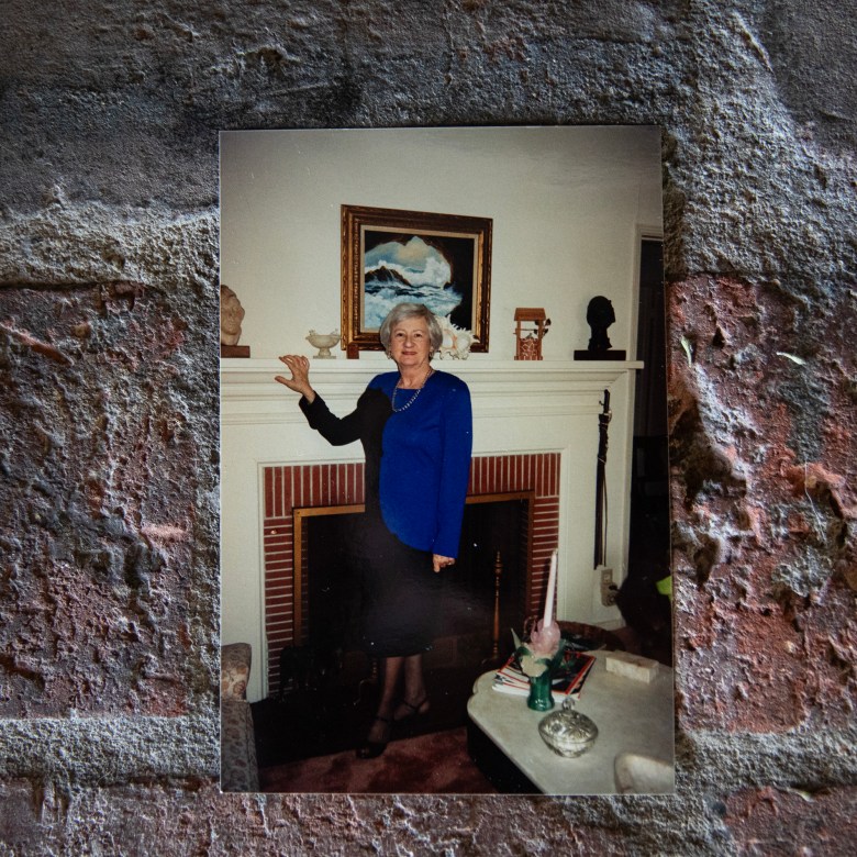 A printed photograph shows an older adult standing beside a fireplace mantel, wearing a blue top and dark skirt, posed in a living room with decorative objects and a framed painting behind them, the print resting against a textured brick surface.