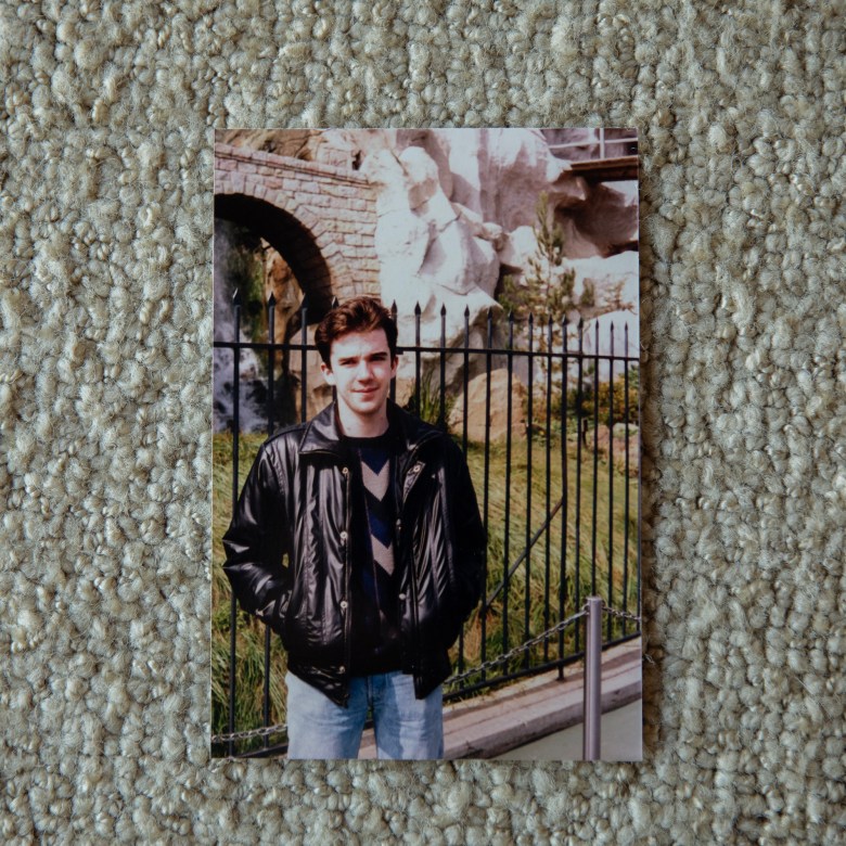 A printed photograph shows a young person in a black leather jacket and jeans standing with hands in pockets in front of a black metal fence, with large rocks and a stone bridge behind them, the print placed on a textured beige carpet.
