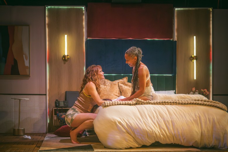 A white women kneels by a hotel bed, clasping the hands of a Black woman sitting on the bed.