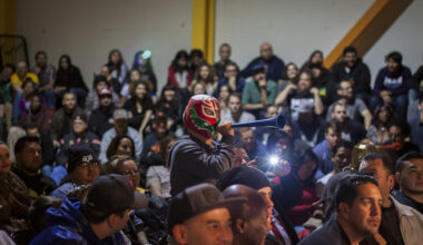 A person in a mask blows a vuvuzela in a crowded indoor audience while others watch an event.