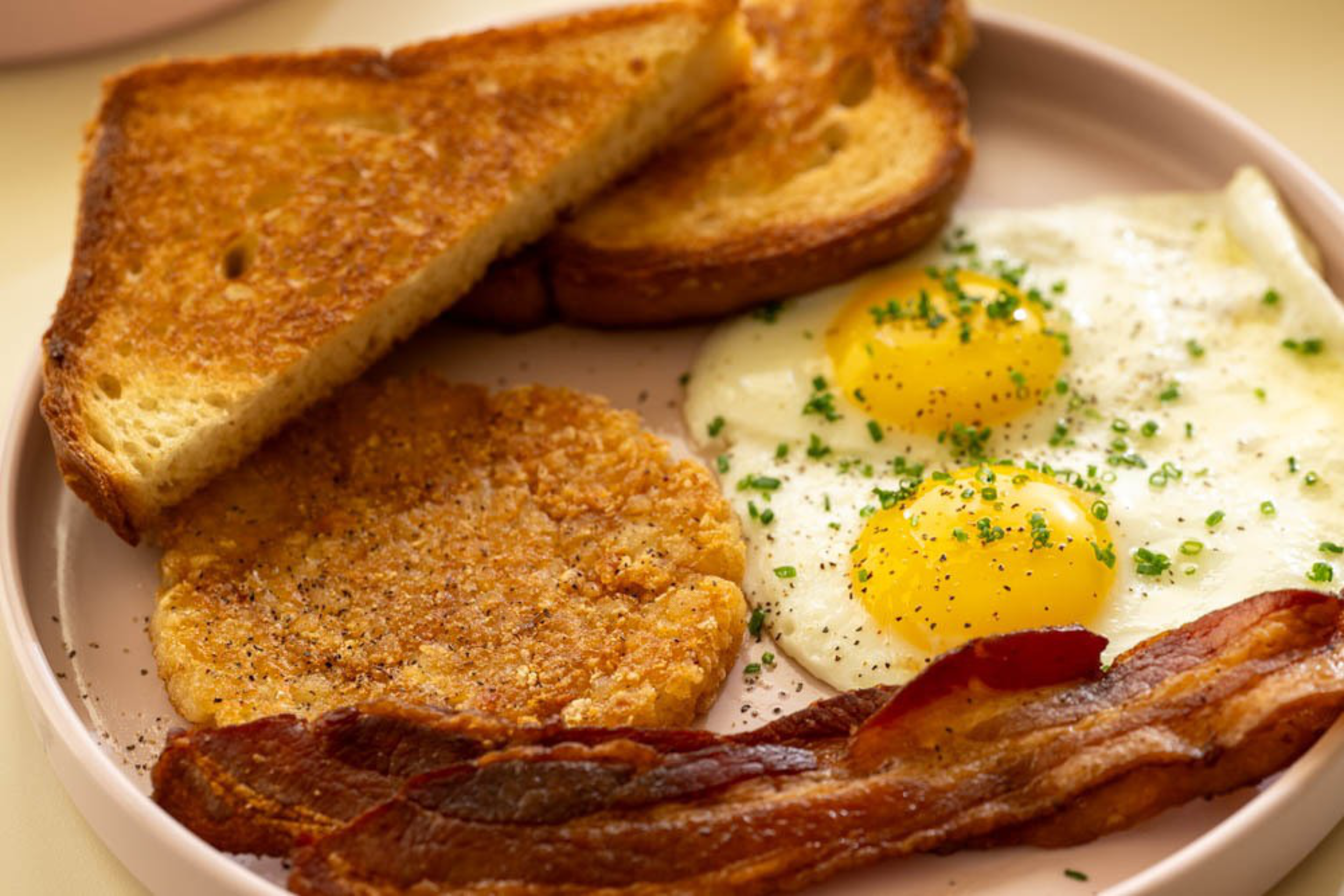 Two sunny-side-up eggs, two crispy bacon strips, a golden hash brown, and two triangular slices of toasted bread on a plate.