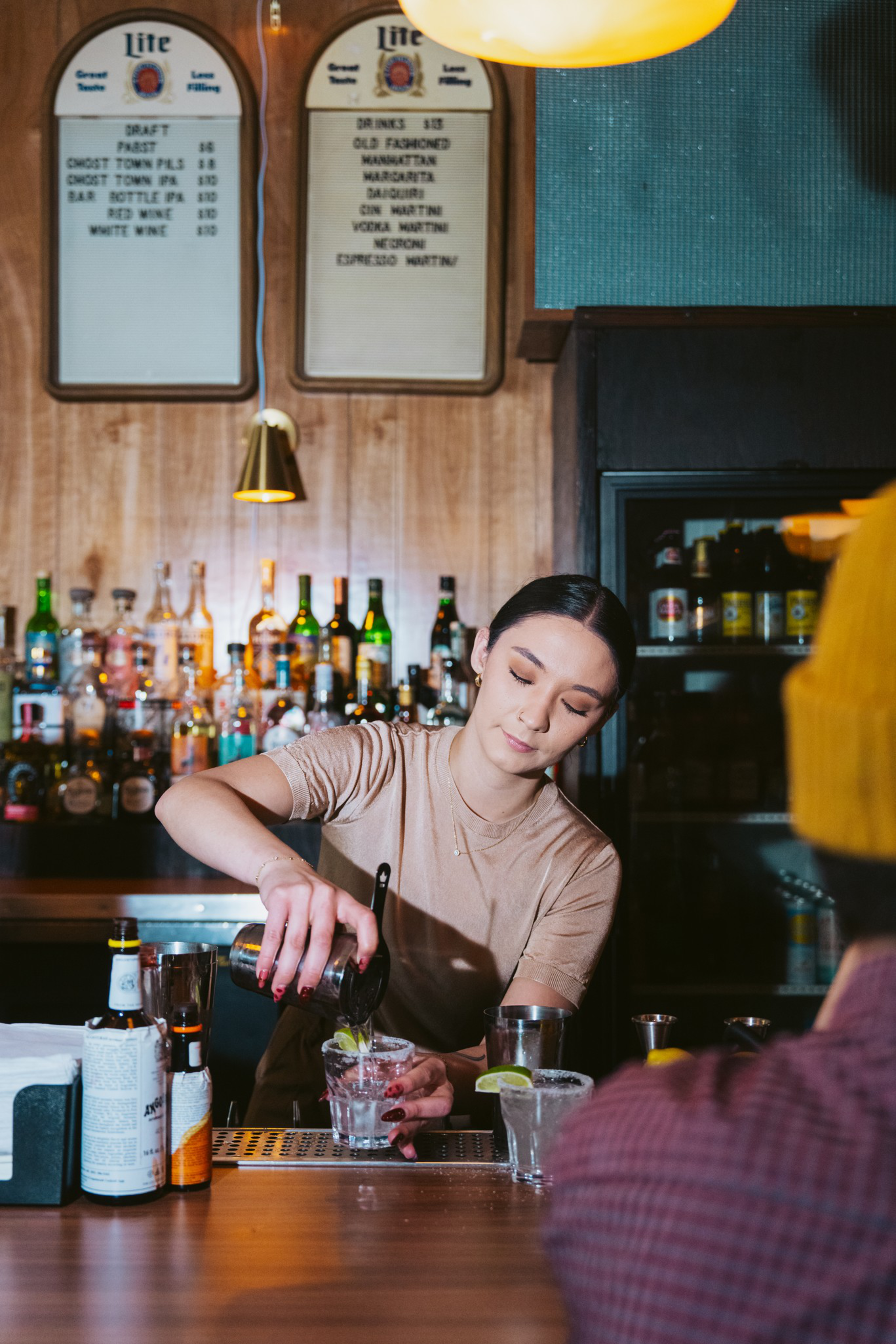 A bartender pours a drink behind a wooden bar, with bottles lined up on shelves and a drink menu on the wall. A customer in a yellow hat sits nearby.