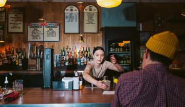 A bartender hands a drink to a customer wearing a yellow beanie inside a dimly lit bar with bottles and menus on the wall behind.