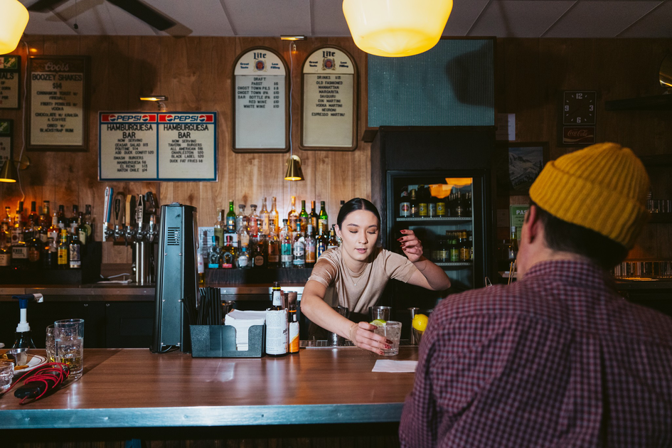 A bartender hands a drink to a customer wearing a yellow beanie inside a dimly lit bar with bottles and menus on the wall behind.