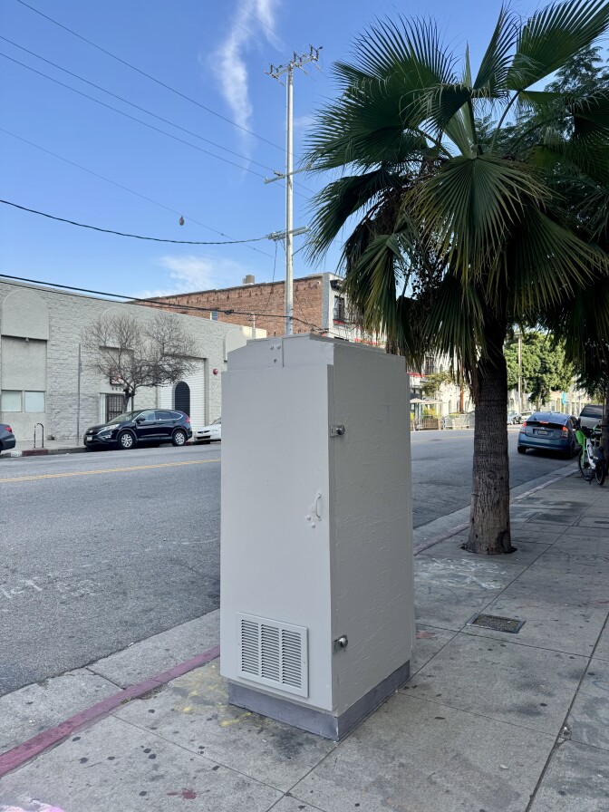 A gray utility box stands closed on a sidewalk near a palm tree and parked cars.