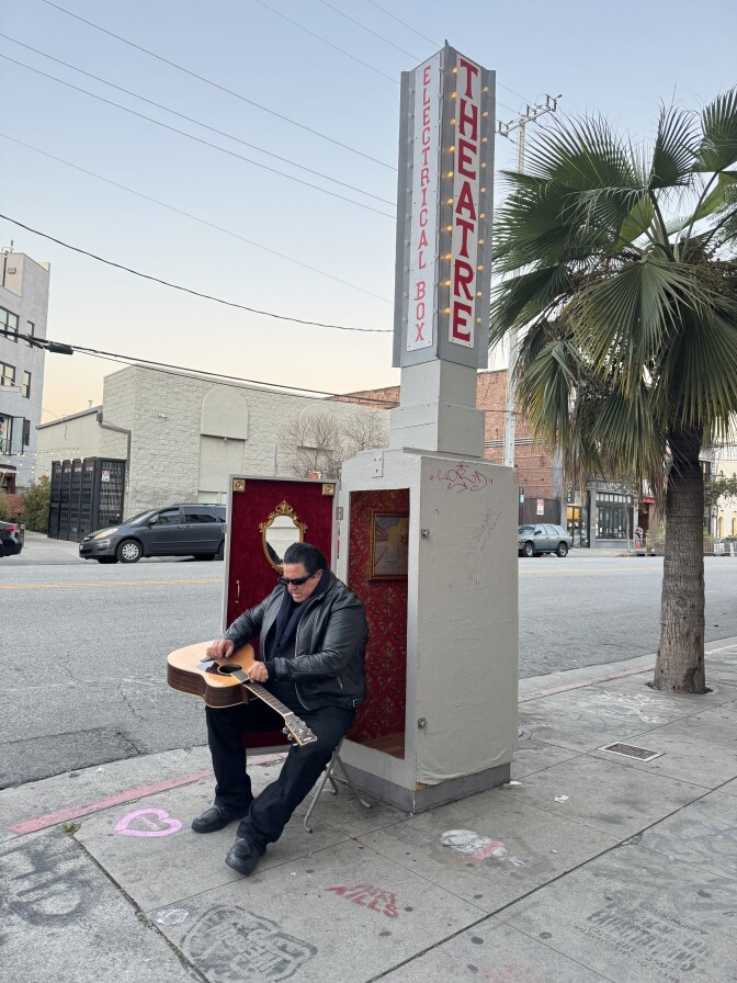 A man in a black jacket sits on an open utility box, tuning a guitar in front of the red velvet-lined interior beneath a lit “Electrical Box Theatre” sign.