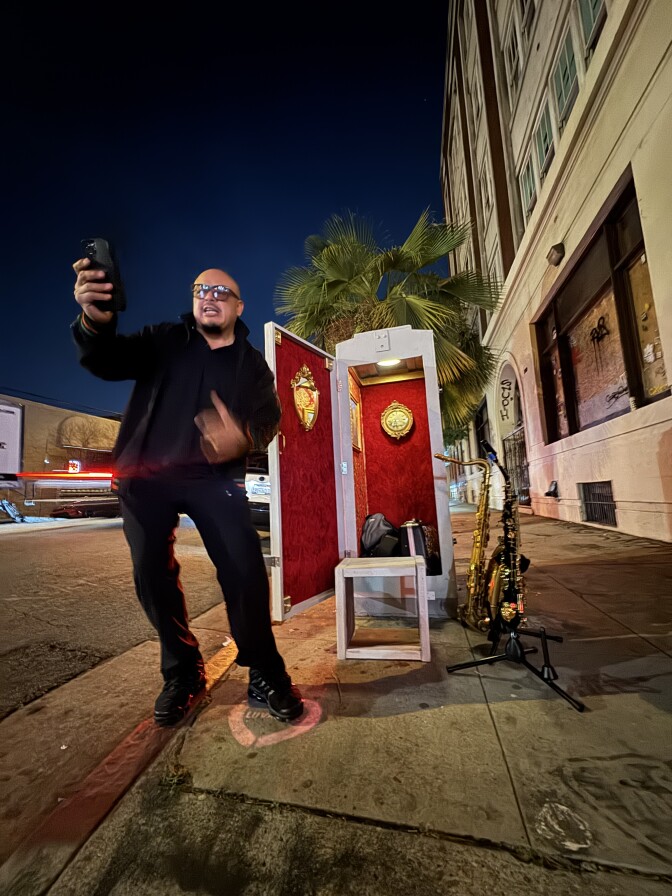 A man gestures while looking at a phone by an open utility box theater with red velvet walls, as two saxophones rest on stands nearby at night.