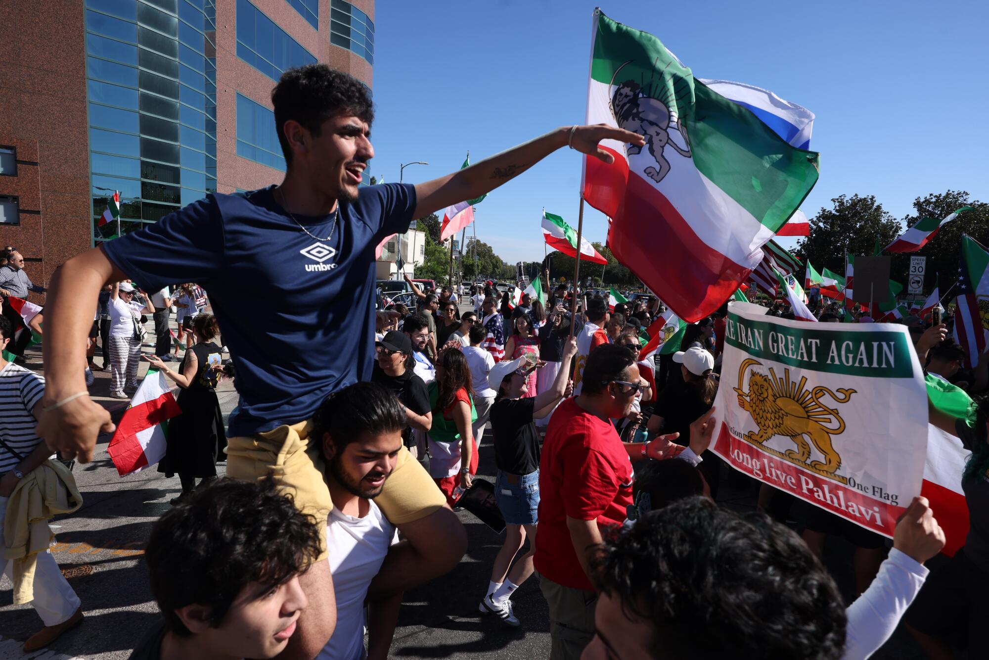 A young man sits on his friend's shoulders at a rally with Iranian flags.