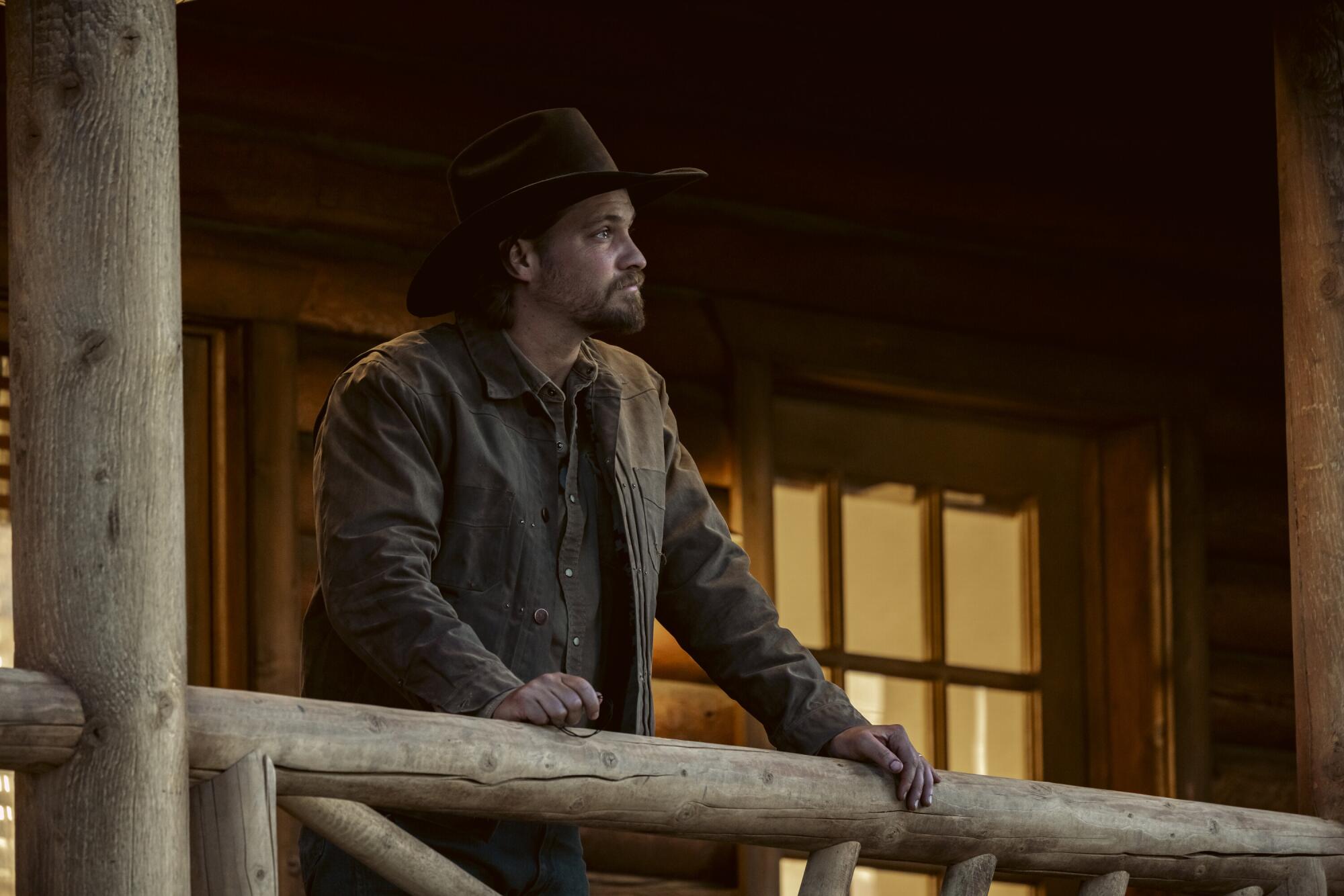 A man in a cowboy hat leans his hands against the railing of the porch of a rustic cabin.