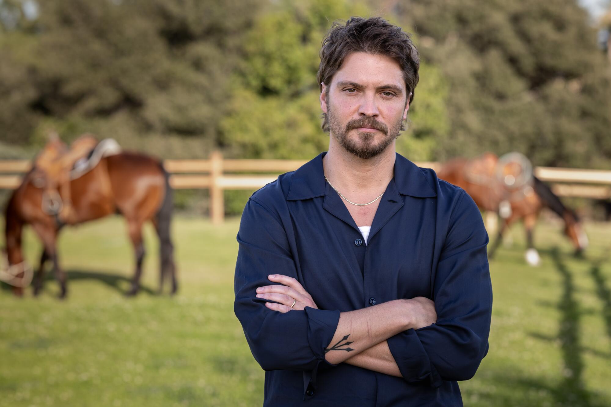 A man in a blue shirt standing with his arms crossed as horses with saddles graze in the background.