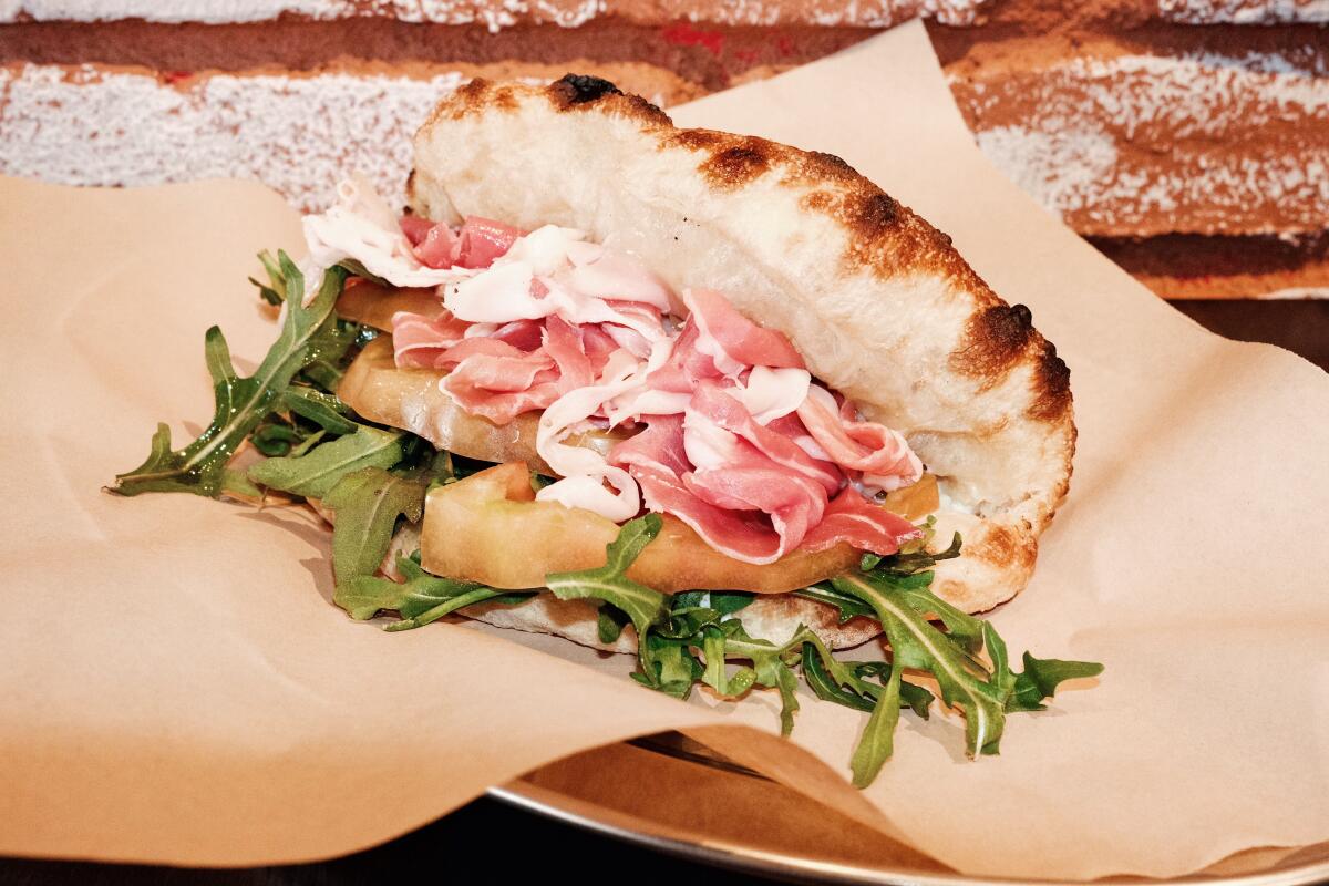 A foldover prosciutto sandwich with stracciatella, heirloom tomato and wild arugula against a brick wall