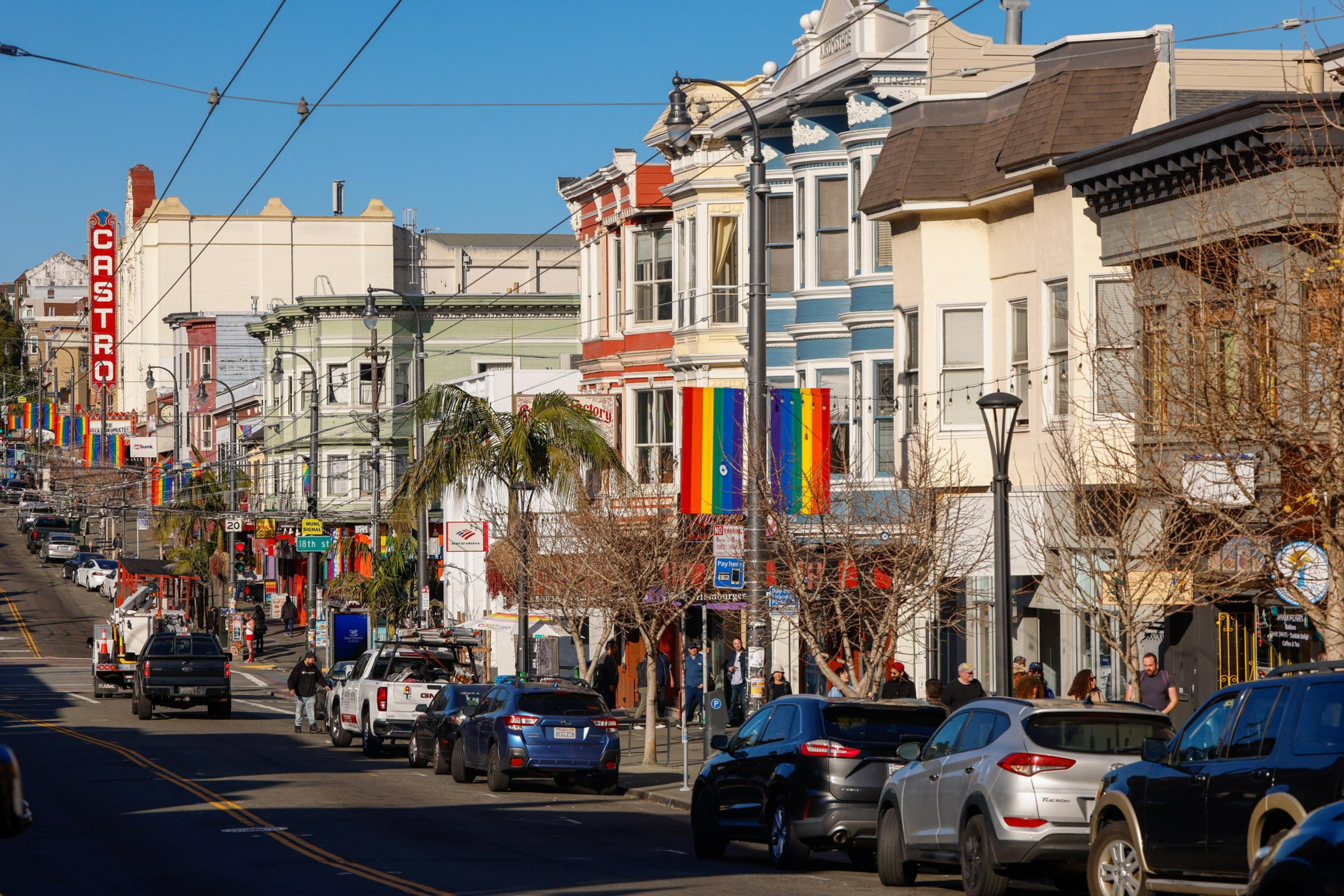 A lively street lined with colorful buildings and rainbow flags, full of parked cars and pedestrians. A large â€œCastroâ€ sign is visible in the background.