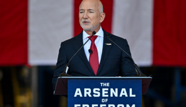 A man in a dark suit and red tie speaks at a podium labeled "The Arsenal of Freedom" with a large American flag in the background.