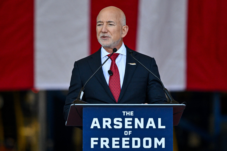 A man in a dark suit and red tie speaks at a podium labeled "The Arsenal of Freedom" with a large American flag in the background.