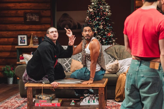 A light-skinned man and a medium-dark-skin-toned man sit on a coffee table, each holding one hand up. The back of a man wearing a red shirt and jeans is to the right of the image.